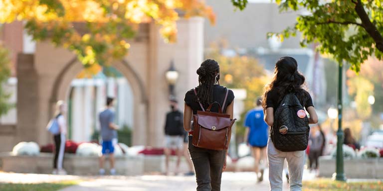The backs of two female students with backpacks walking towards the Sample Gates.